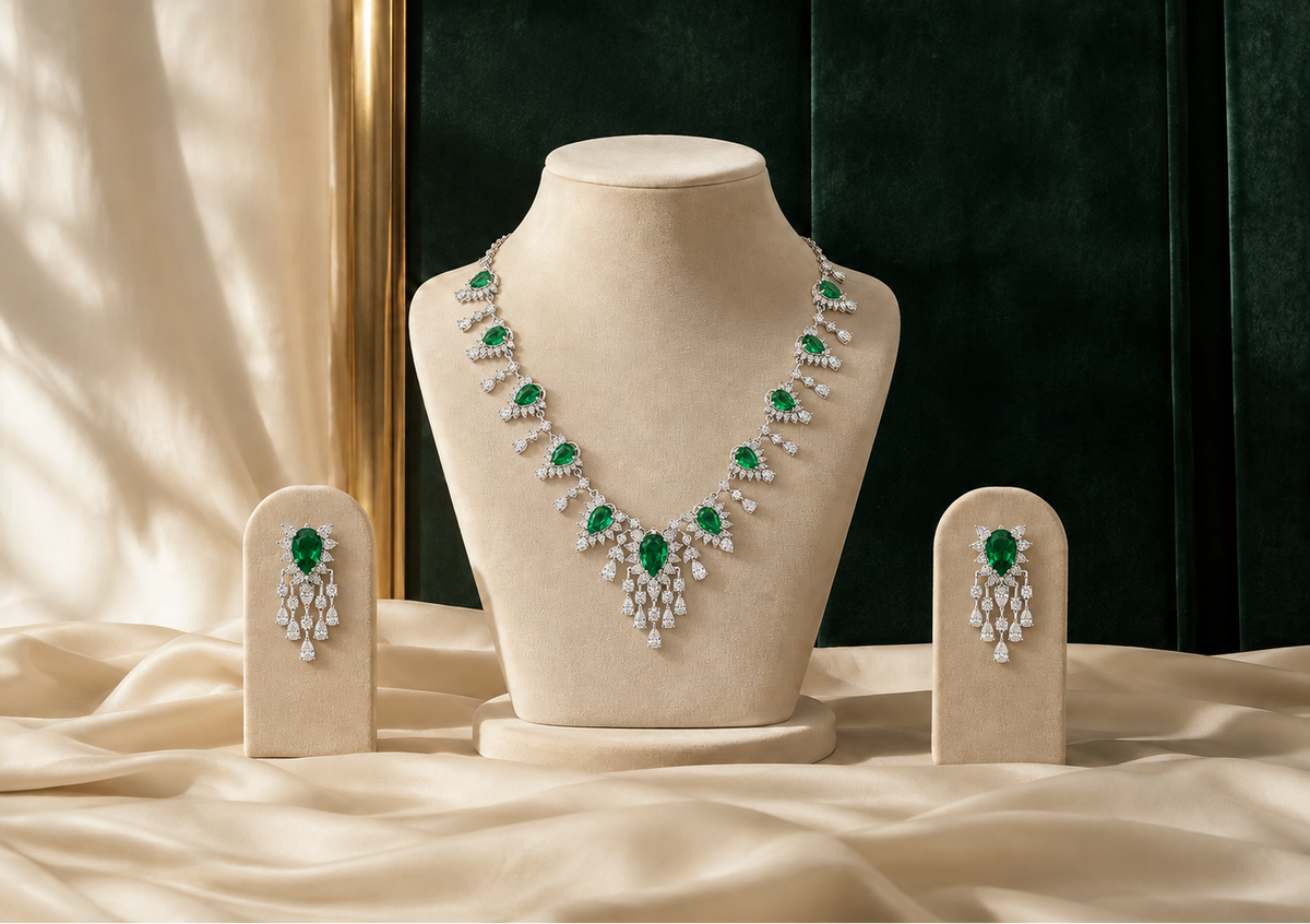 A luxury jewelry set featuring a necklace and matching drop earrings displayed on beige velvet mannequin stands. The jewelry is set with vivid green emeralds and brilliant white diamonds, arranged on draped champagne silk fabric against a dark green velvet background.